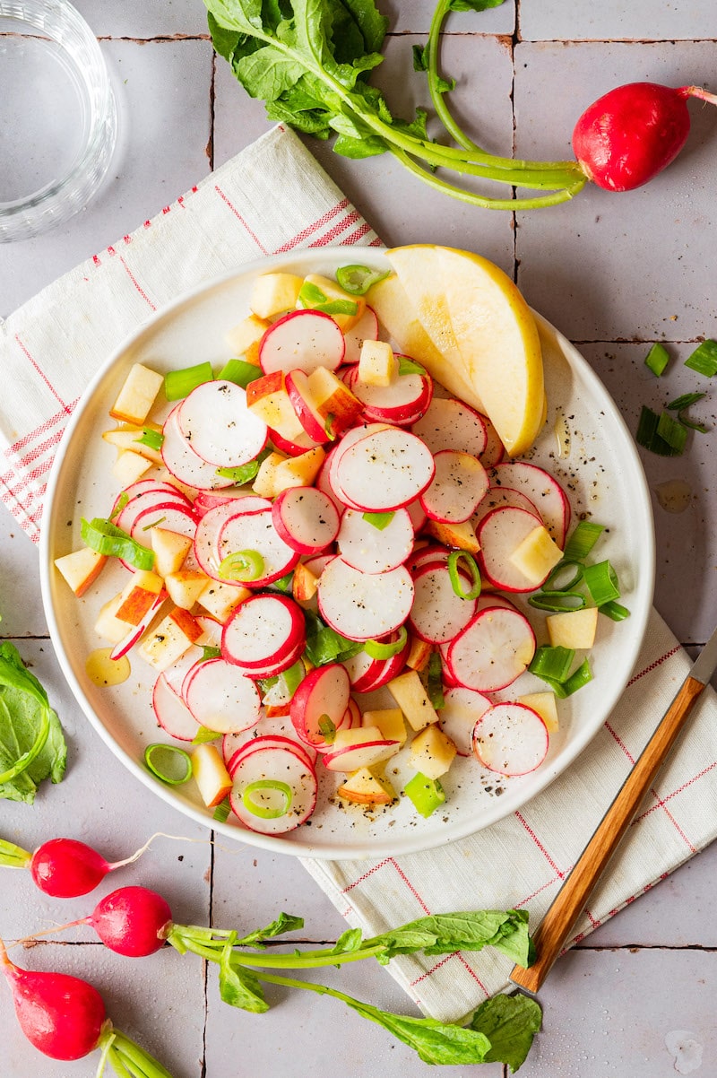 Apfel-Radieschen-Salat mit Frühlingszwiebeln. Um den Teller herum sind frische Radieschen dekoriert.