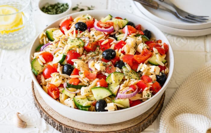 Mediterraner Kritharaki-Salat mit Nudelreis, Gurken, Tomaten, Paprika, Oliven und roten Zwiebeln in einer weißen Schüssel serviert.