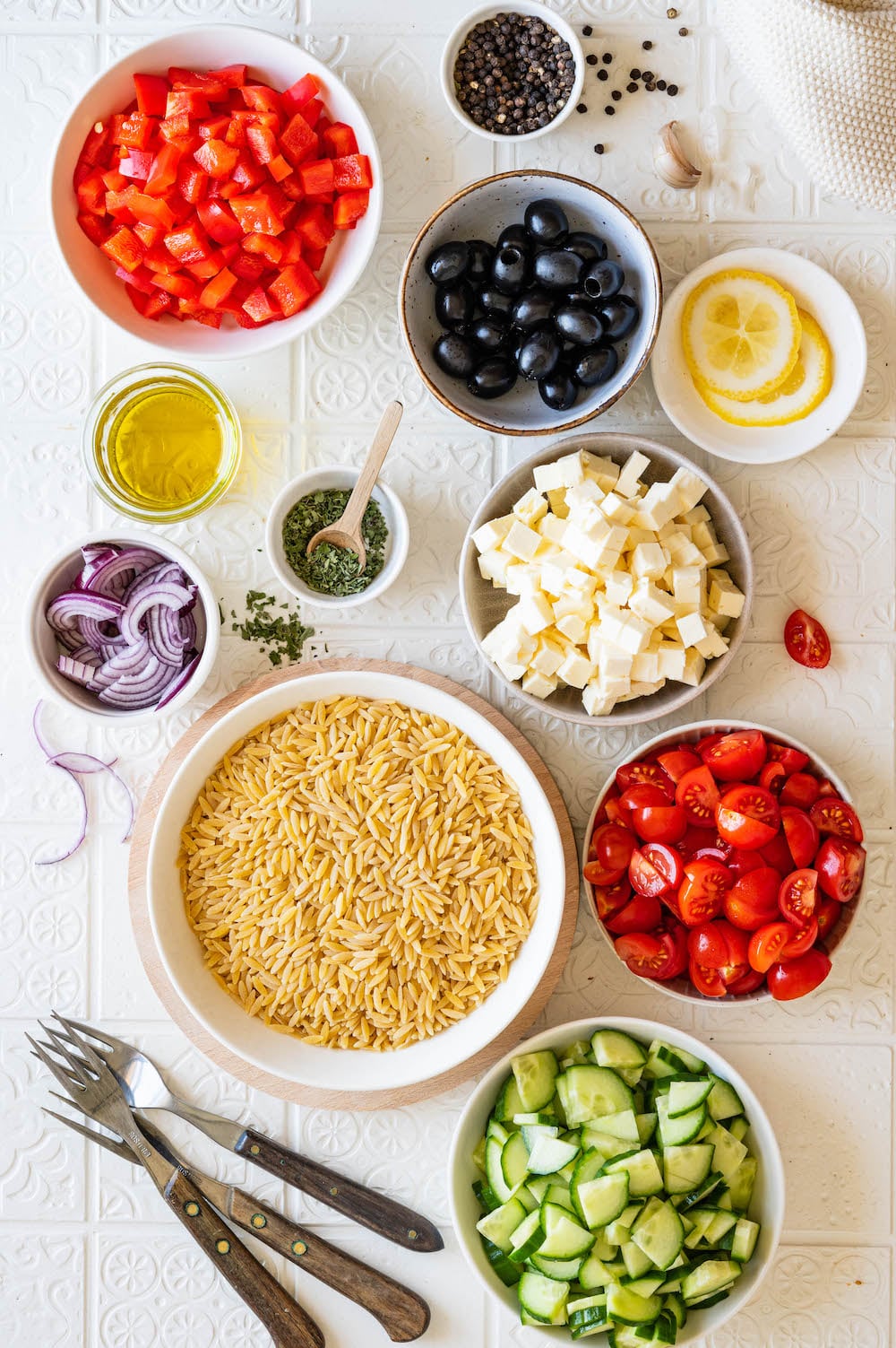 Zutaten für den Kritharaki-Salat in Schüsselchen von oben fotografiert: Kritharaki, Gurke, rote Paprika, Tomaten, rote Zwiebel, schwarze Oliven, Knoblauch, Olivenöl und Oregano.