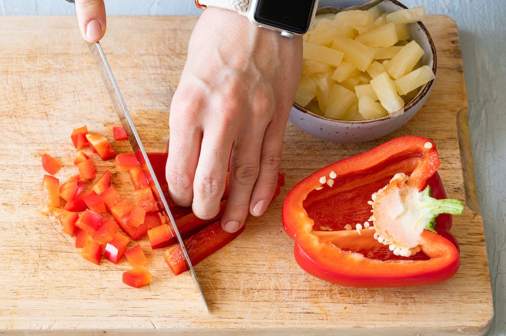 Paprika wird in kleine Würfel geschnitten. Daneben stehen Ananas in einer Schüssel.