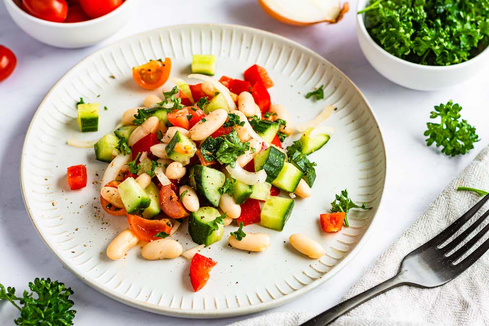 Salat mit weißen Bohnen, Gurken, Tomaten und Paprika auf einem Teller serviert, im Hintergrund sind die einzelnen Zutaten nochmal dekoriert.