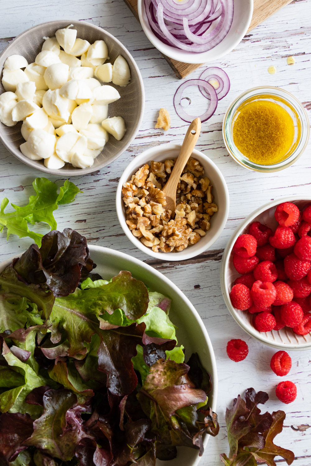 Zutaten für den Himbeersalat in Schüsseln: Blattsalat, Mozzarella, Walnüsse, Himbeeren, rote Zwiebeln und ein Dressing in einem Glas.