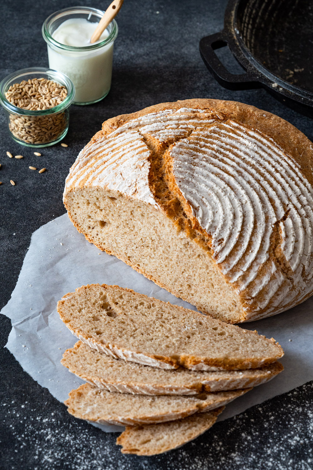 Joghurt-Dinkel-Brot mit Kruste auf dunklem Untergrund. Vier Scheiben sind vom Laib abgeschnitten und liegen aufgefächert vor dem Brot.