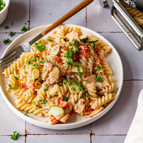 Pasta mit Lachs, Zucchini und Tomaten auf einem flachen Teller serviert und mit Petersilie bestreut.