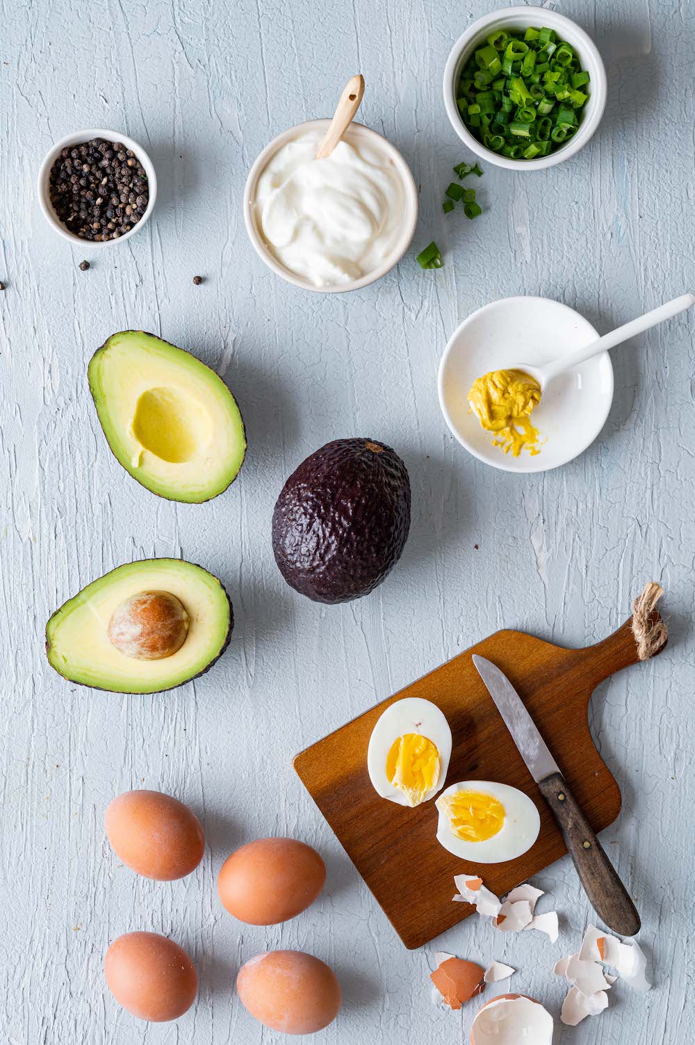 Zutaten für den Avocado-Eier Salat: Hart gekochte Eier, reife Avocado, Joghurt, Senf, Frühlingszwiebeln und Pfeffer.